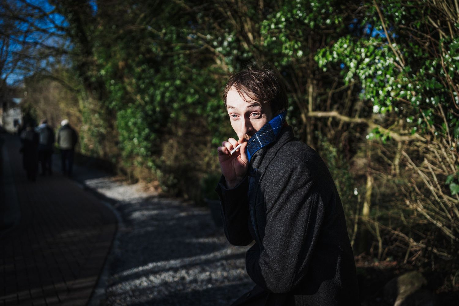 A person in a black coat stands on a shadowy path surrounded by greenery while making a quiet gesture.