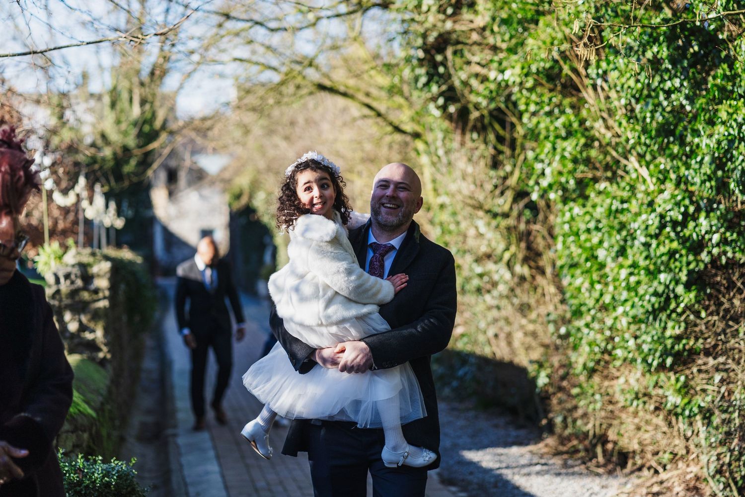 A joyful embrace between two people on a sunny path lined with greenery and trees.