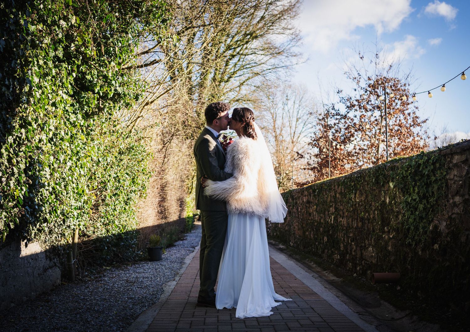 A couple embraces on a tree-lined path with stone walls on a sunny autumn day.