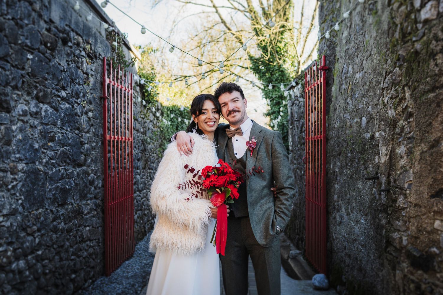 A couple poses between stone walls holding a vibrant red bouquet on their wedding day.