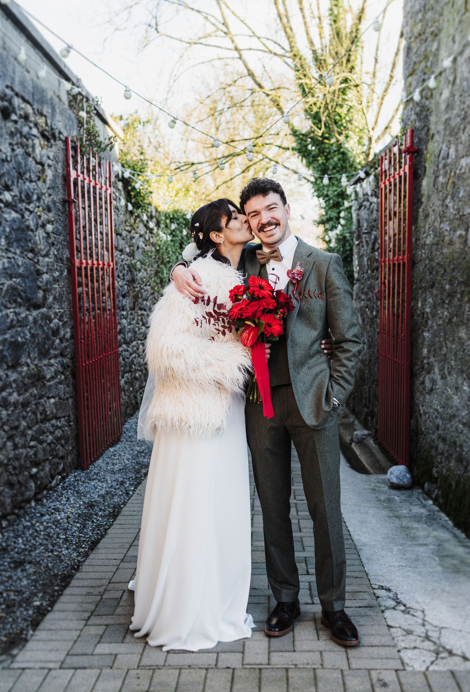 Wedding couple posing by stone walls and red gate in an outdoor garden setting.