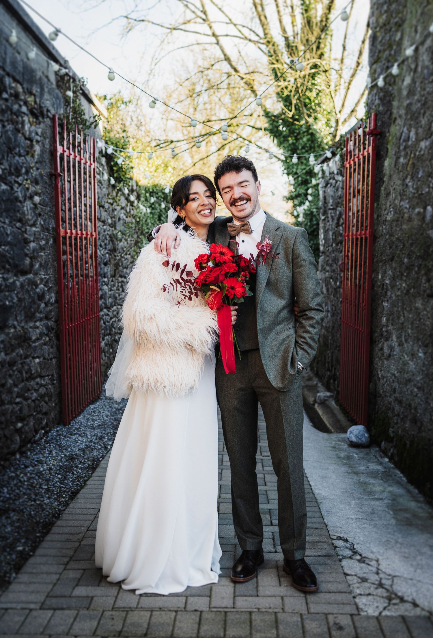 A joyful couple shares candid moments in their wedding attire along a stone-walled pathway with red gates.