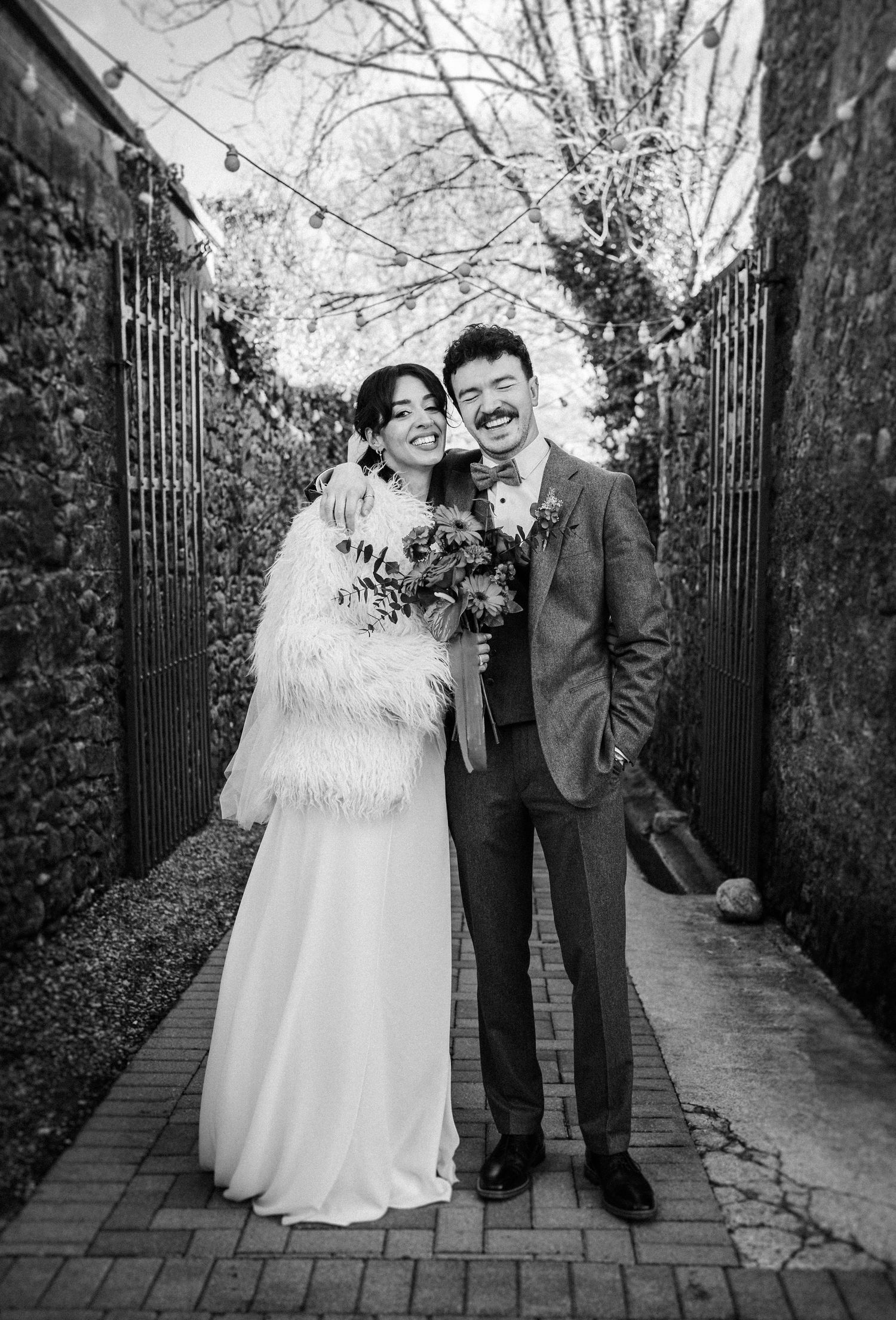 A wedding couple shares joyful moments together on a brick path surrounded by greenery in black and white photography.