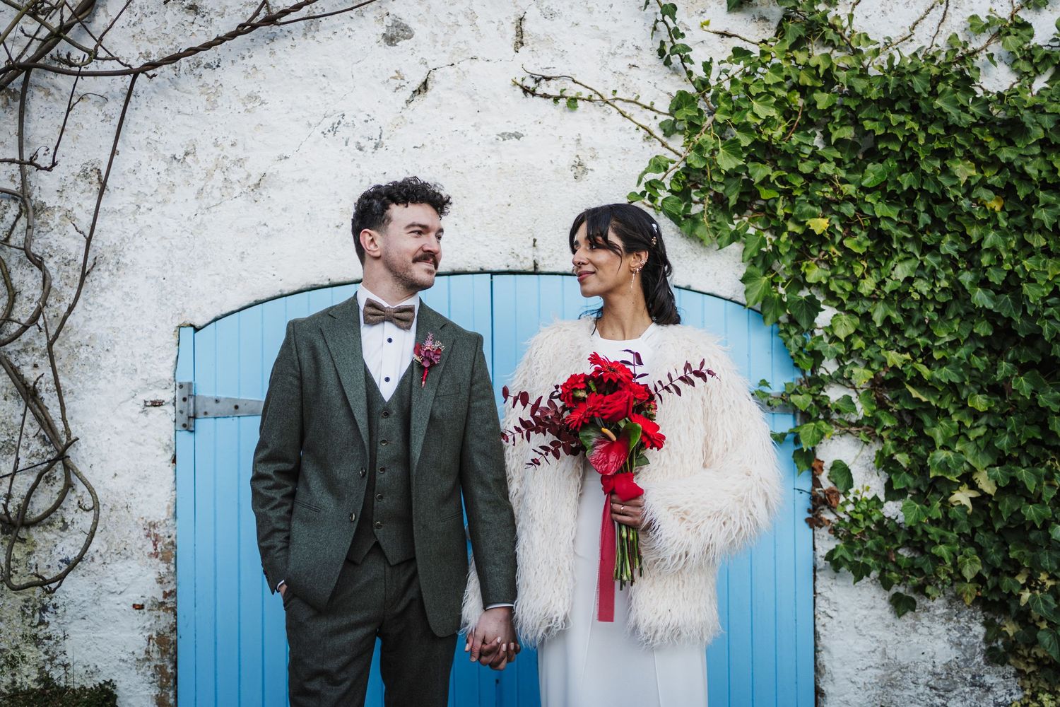 Newlywed couple poses against rustic white wall with blue door and climbing ivy, bride holding red bouquet.