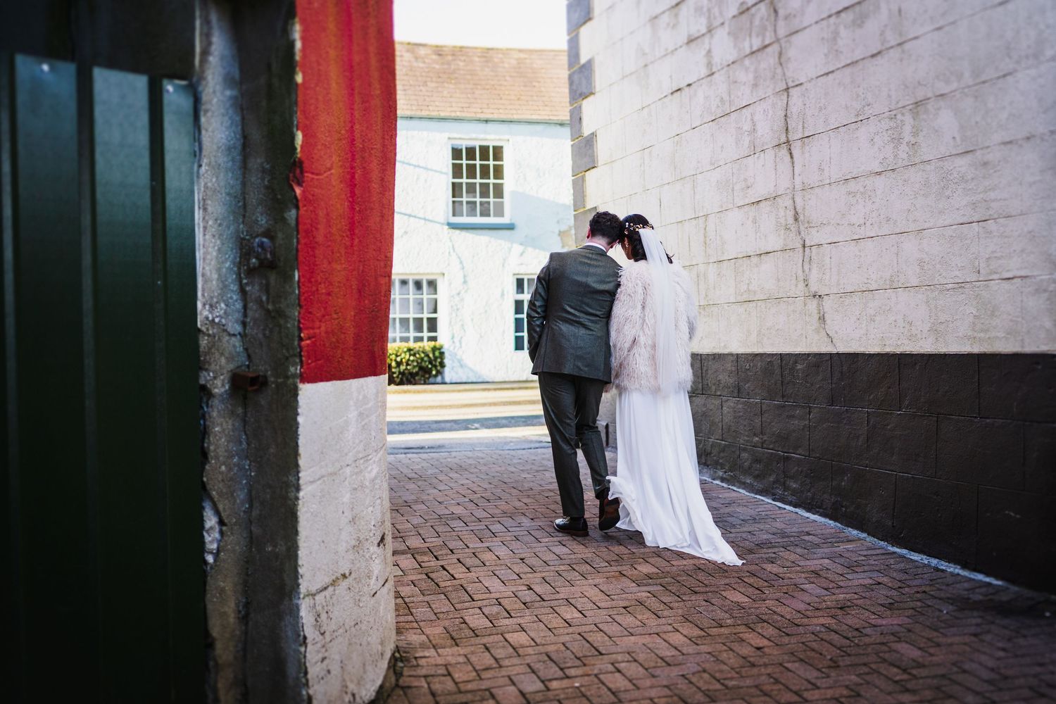 A couple in wedding attire shares an intimate moment in a brick alleyway with white colonial buildings in the background.