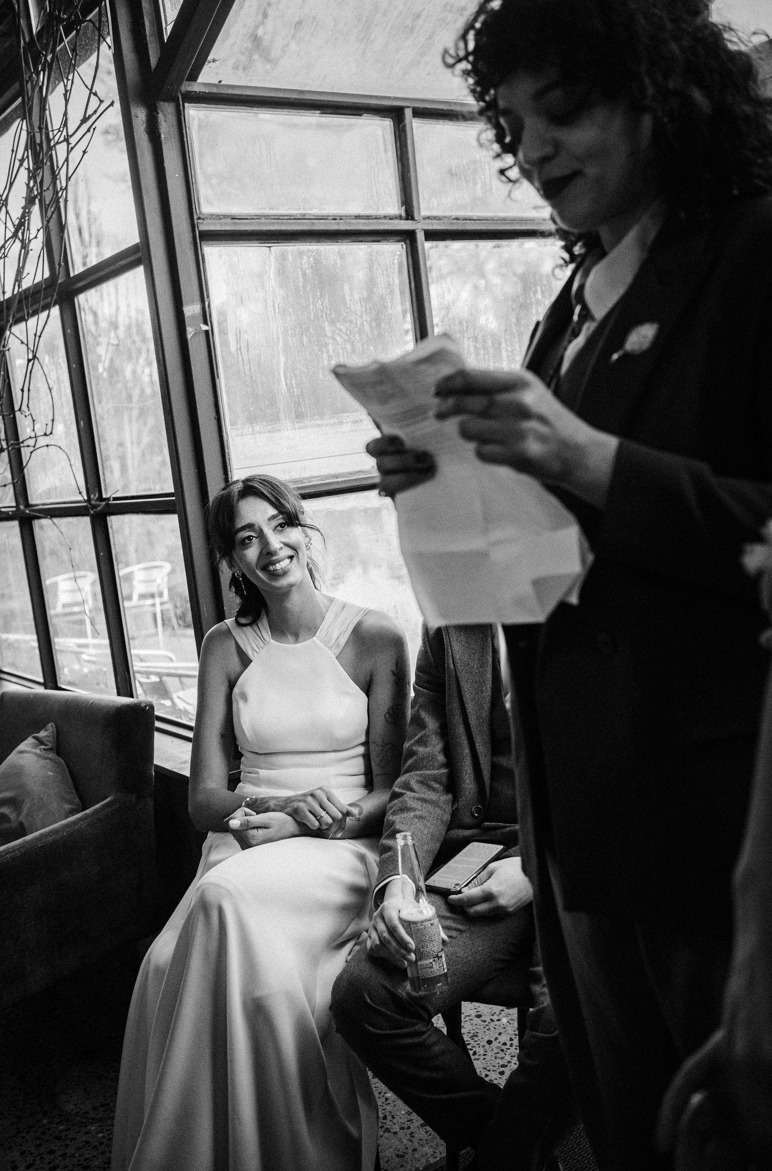 A black and white wedding photo showing a bride in a white dress seated by a window while someone reads from papers.