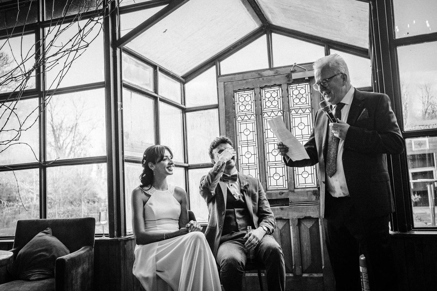 Black and white panoramic photograph of a wedding reception in a glass-walled venue with guests mingling and dancing.