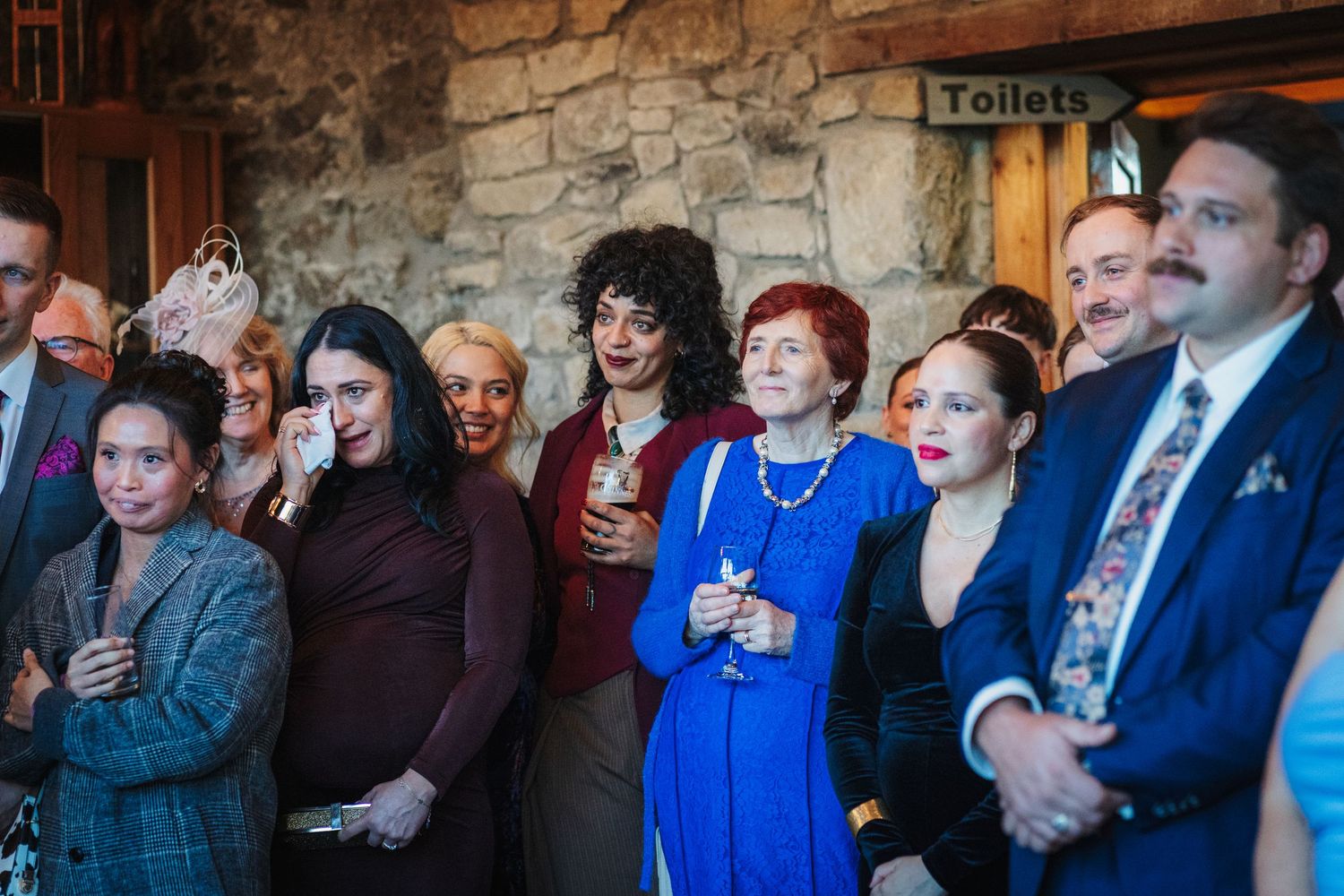 A group of wedding guests stand together against a stone wall during a reception ceremony.