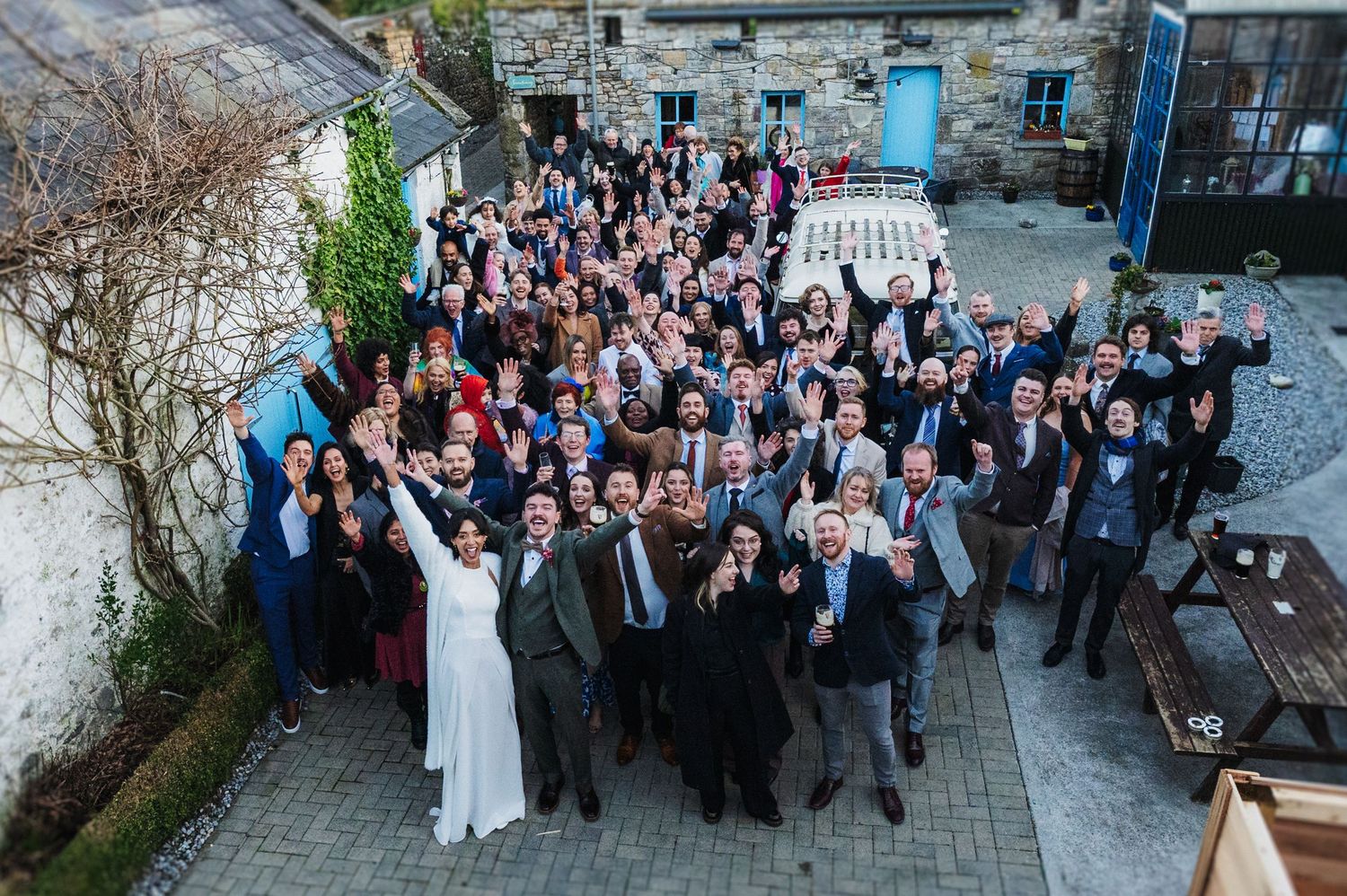 Large wedding party group photo taken from above showing guests gathered in courtyard celebrating with raised arms.