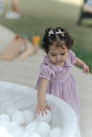 A small child in a purple dress touches and explores fluffy white cotton-like material spread out on a surface.