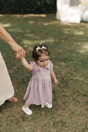 A toddler in a lavender dress takes their first steps on grass while being supported by an adult's hand.