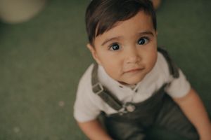 Young toddler in overalls looking up at camera with green carpet background.