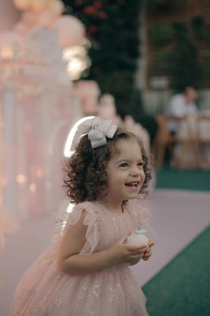A small girl in a pink party dress laughs while holding something at an outdoor celebration.