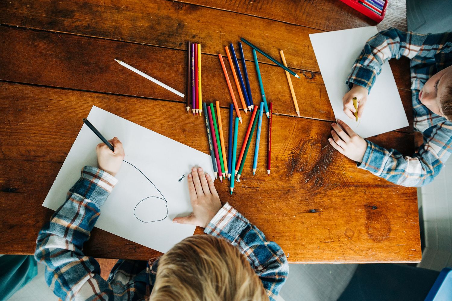 Overhead view of hands writing and drawing with colored pencils and art supplies on a wooden desk surface.