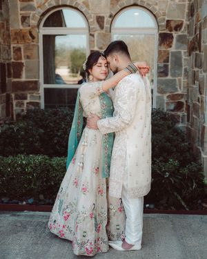 A couple in traditional South Asian wedding attire shares romantic moments against a stone wall backdrop.