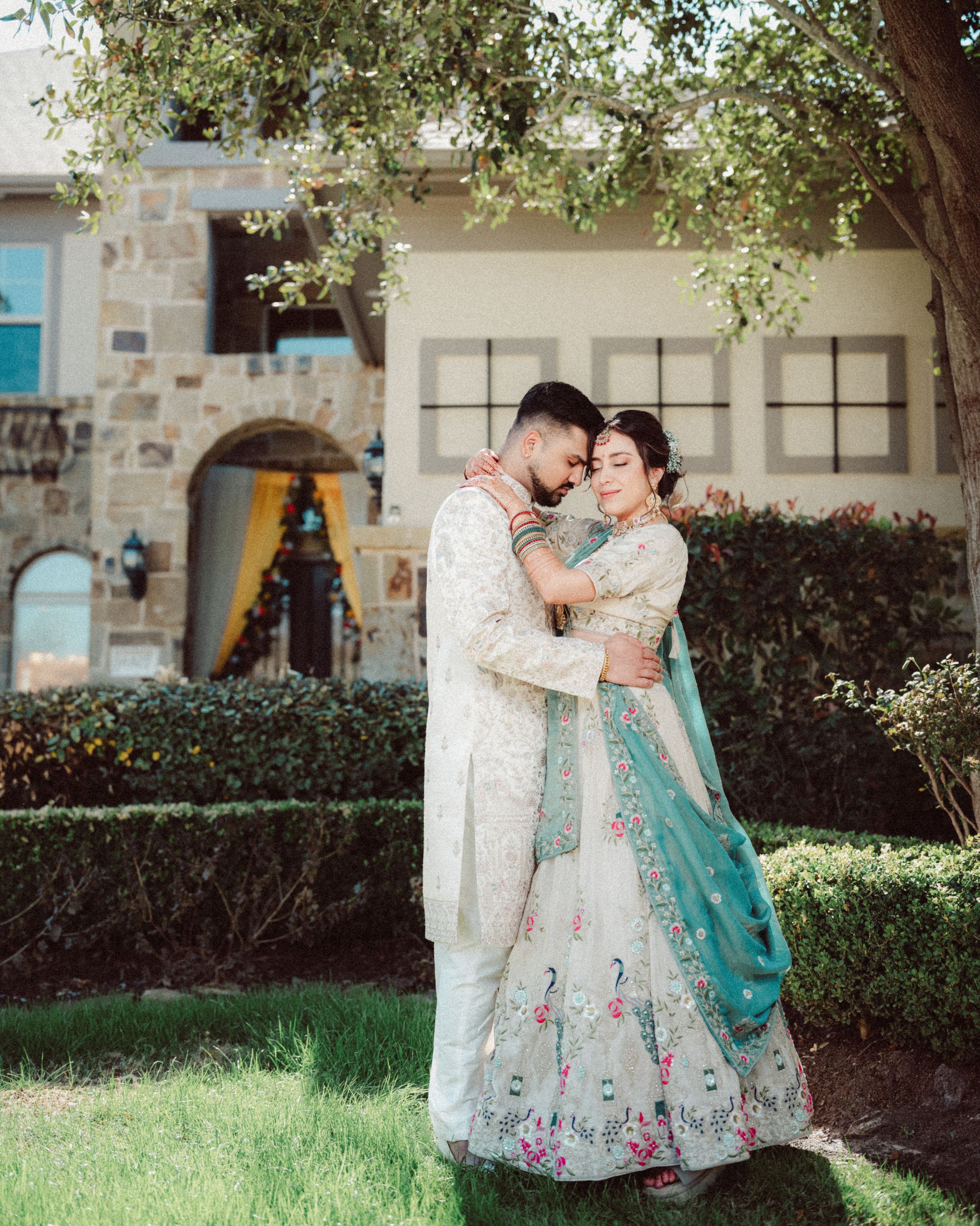 A couple in traditional South Asian wedding attire share romantic moments in a garden setting under trees.