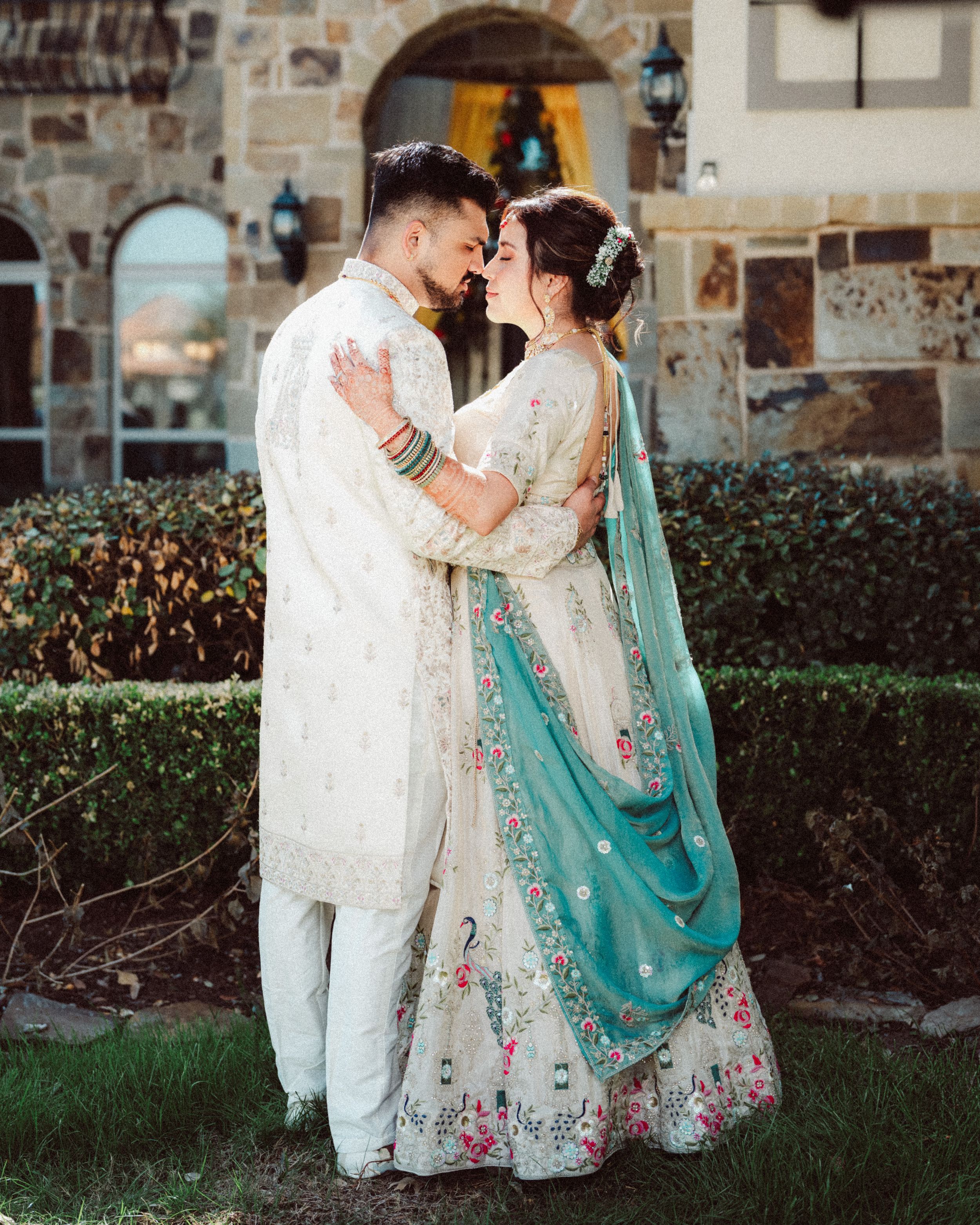A couple in traditional South Asian wedding attire share an intimate moment in a garden setting.