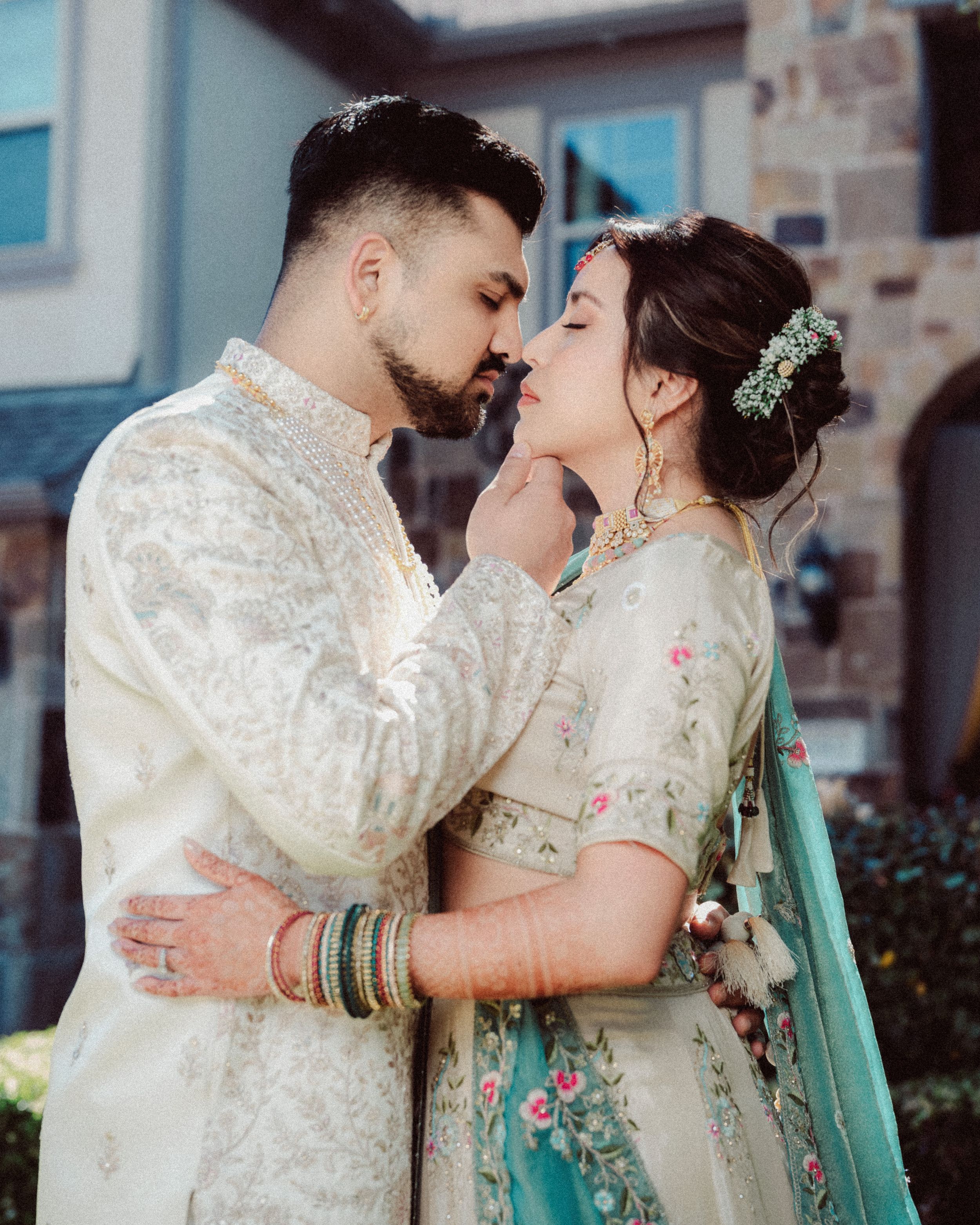 A romantic couple in traditional white and floral wedding attire share an intimate moment outdoors.