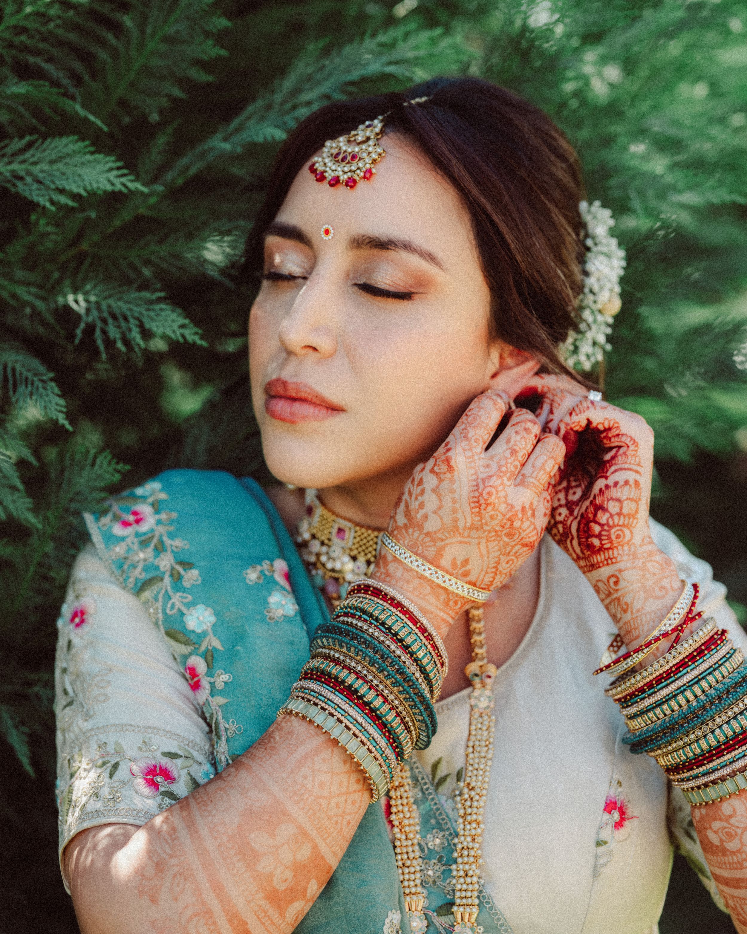 Close up details of intricate henna designs and bangles on hands against a dreamy green outdoor background.