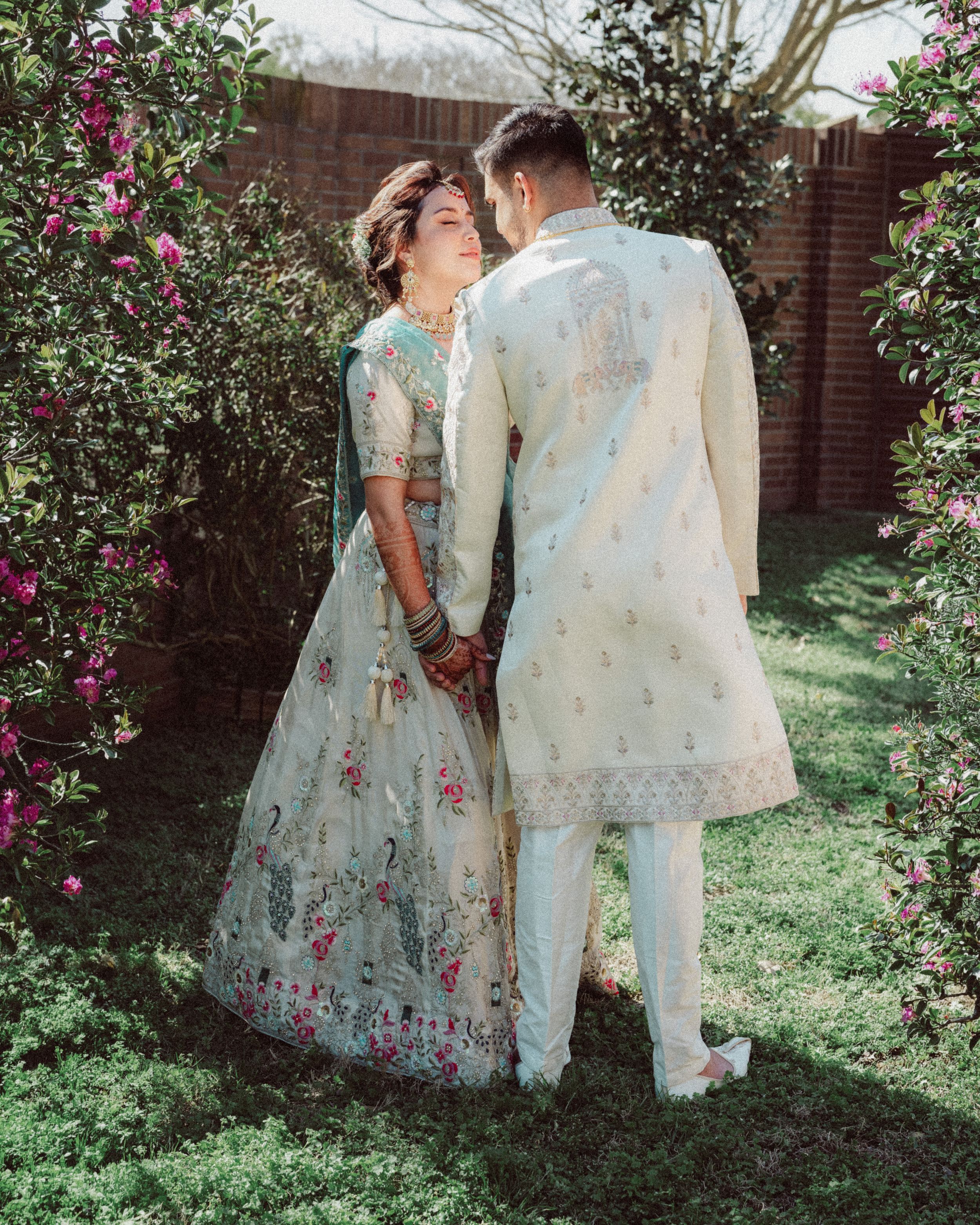 A couple in traditional South Asian wedding attire embrace in a garden with pink flowers and brick wall backdrop.