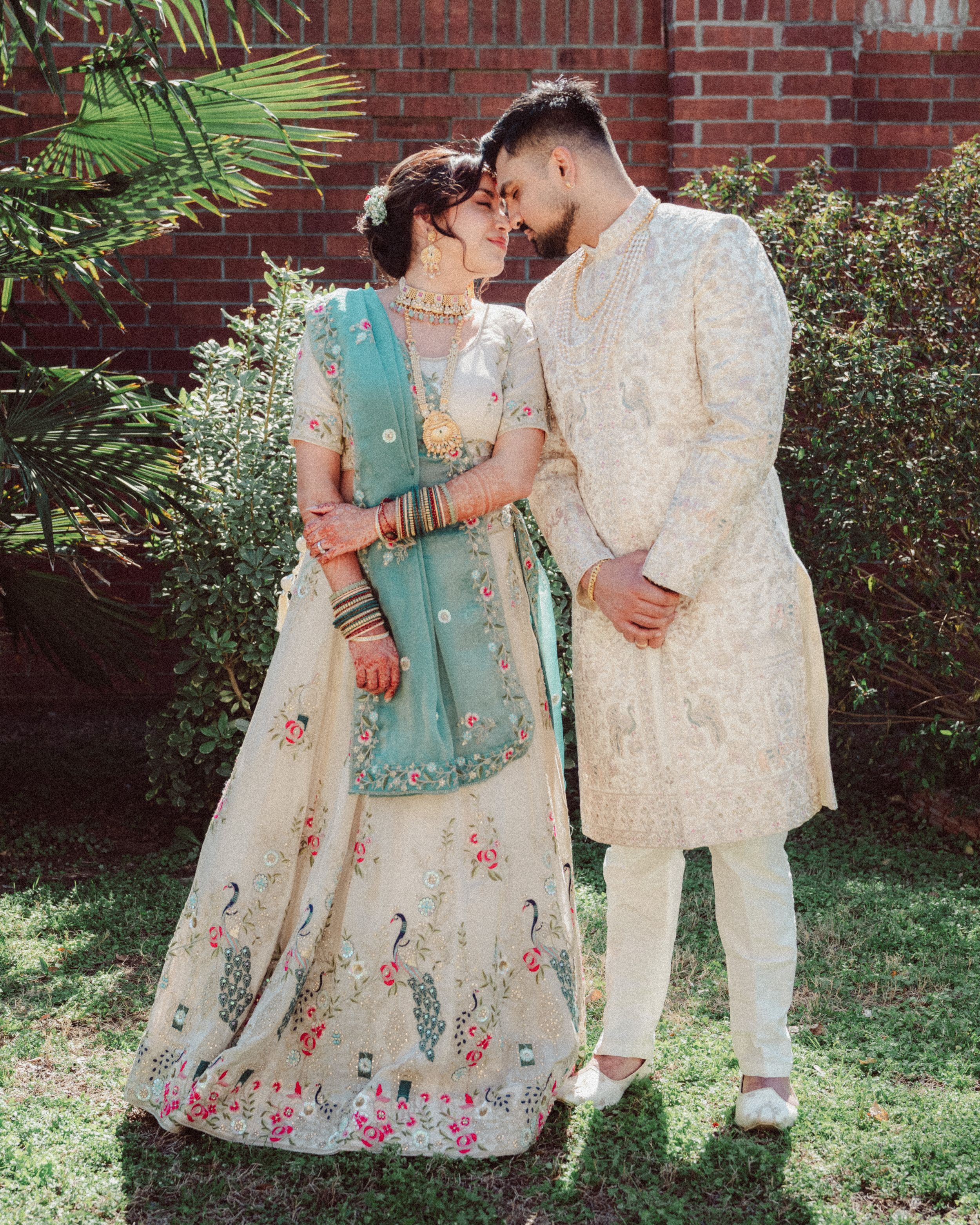 A couple in traditional South Asian attire embraces outdoors against a brick wall and garden backdrop.