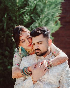 A couple in traditional South Asian wedding attire embrace outdoors against greenery in an intimate portrait.