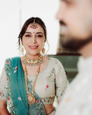 Bride in traditional Indian wedding attire with embroidered teal dupatta and ornate gold jewelry smiles during ceremony.