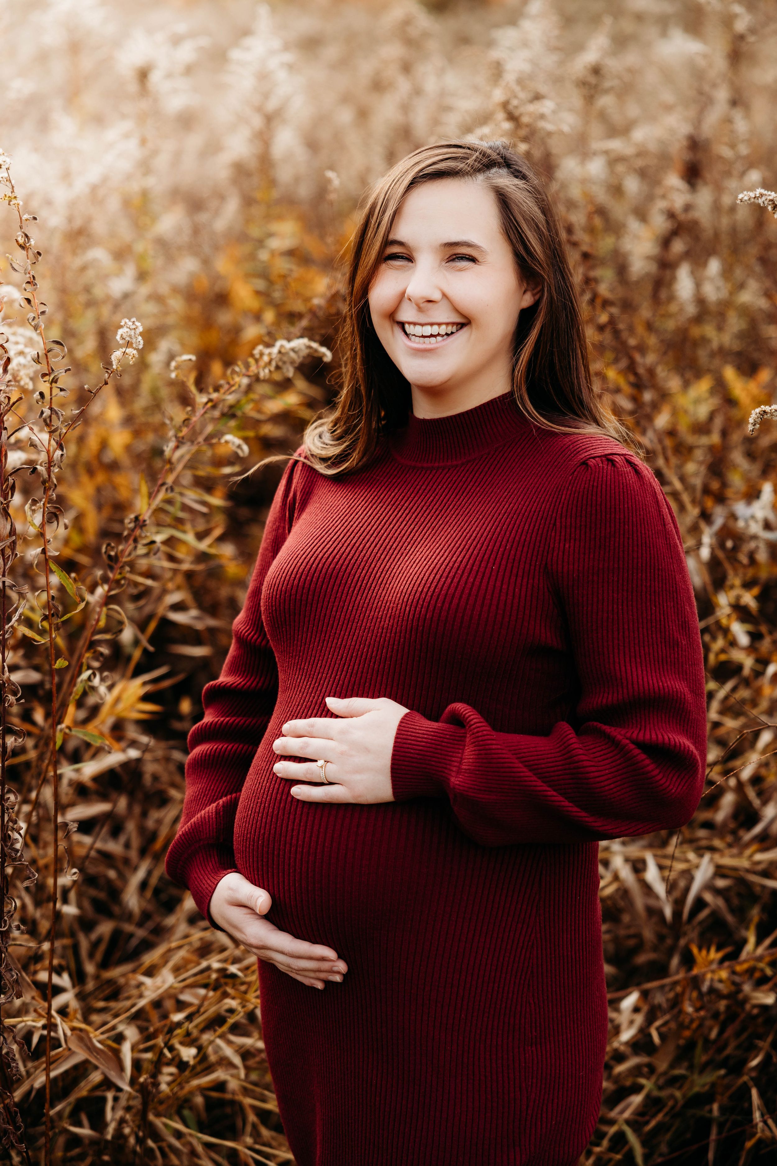 A person in a burgundy sweater dress poses outdoors during autumn while cradling their pregnant belly.