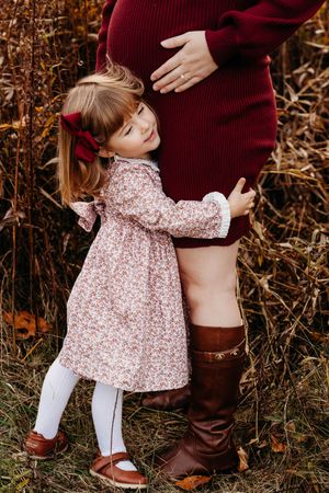 Small child in floral dress hugging someone wearing burgundy sweater and brown boots in autumn setting.