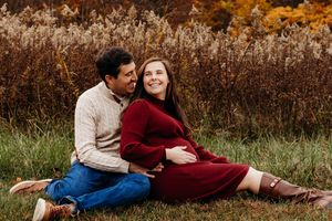 Couple in casual sweaters sitting together in autumn field with golden brown foliage and tall grass in background.