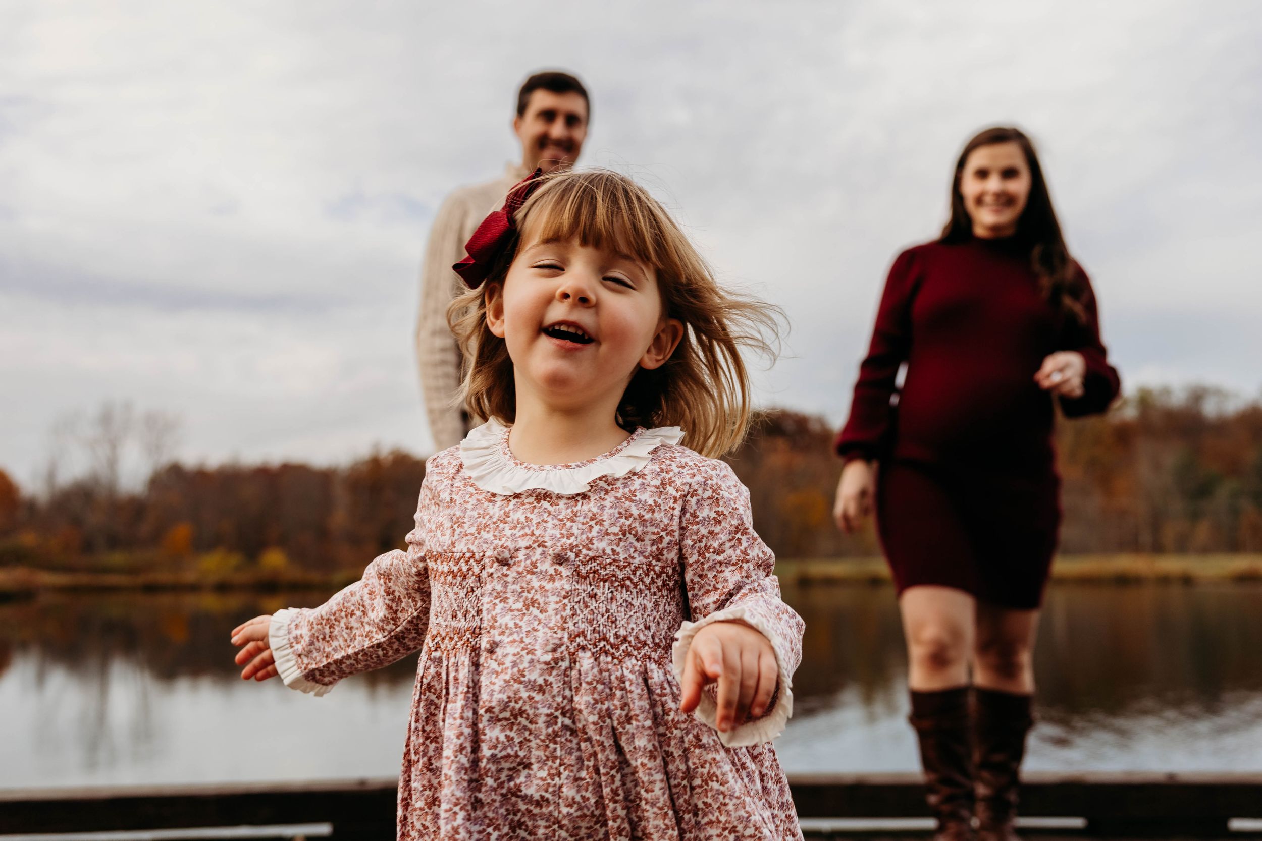 A young child in a floral dress runs joyfully on a dock by a lake during autumn with others in the background.