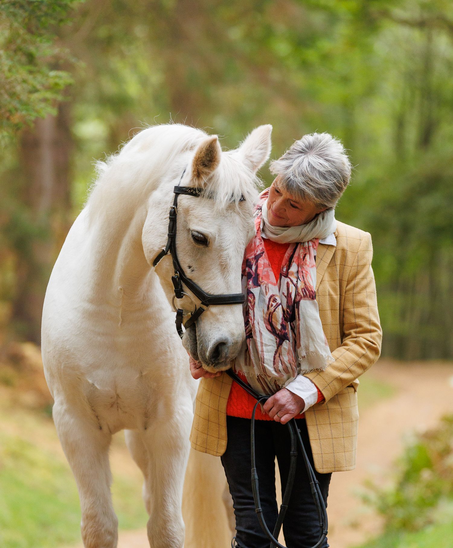 A brown horse and rider in a white sweater pose together against autumn foliage in soft natural lighting.