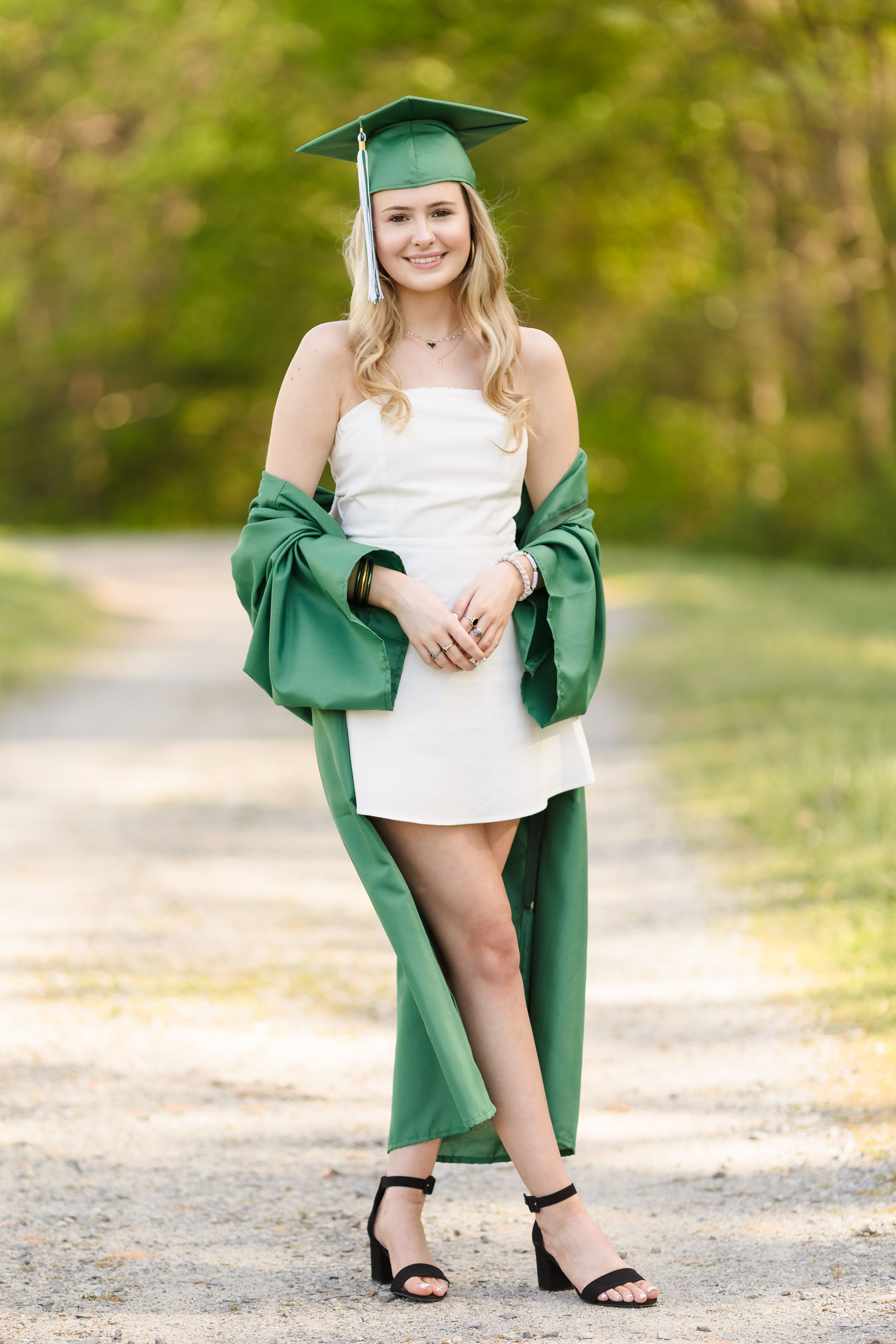 Graduate in green cap and gown with white dress poses on outdoor path.