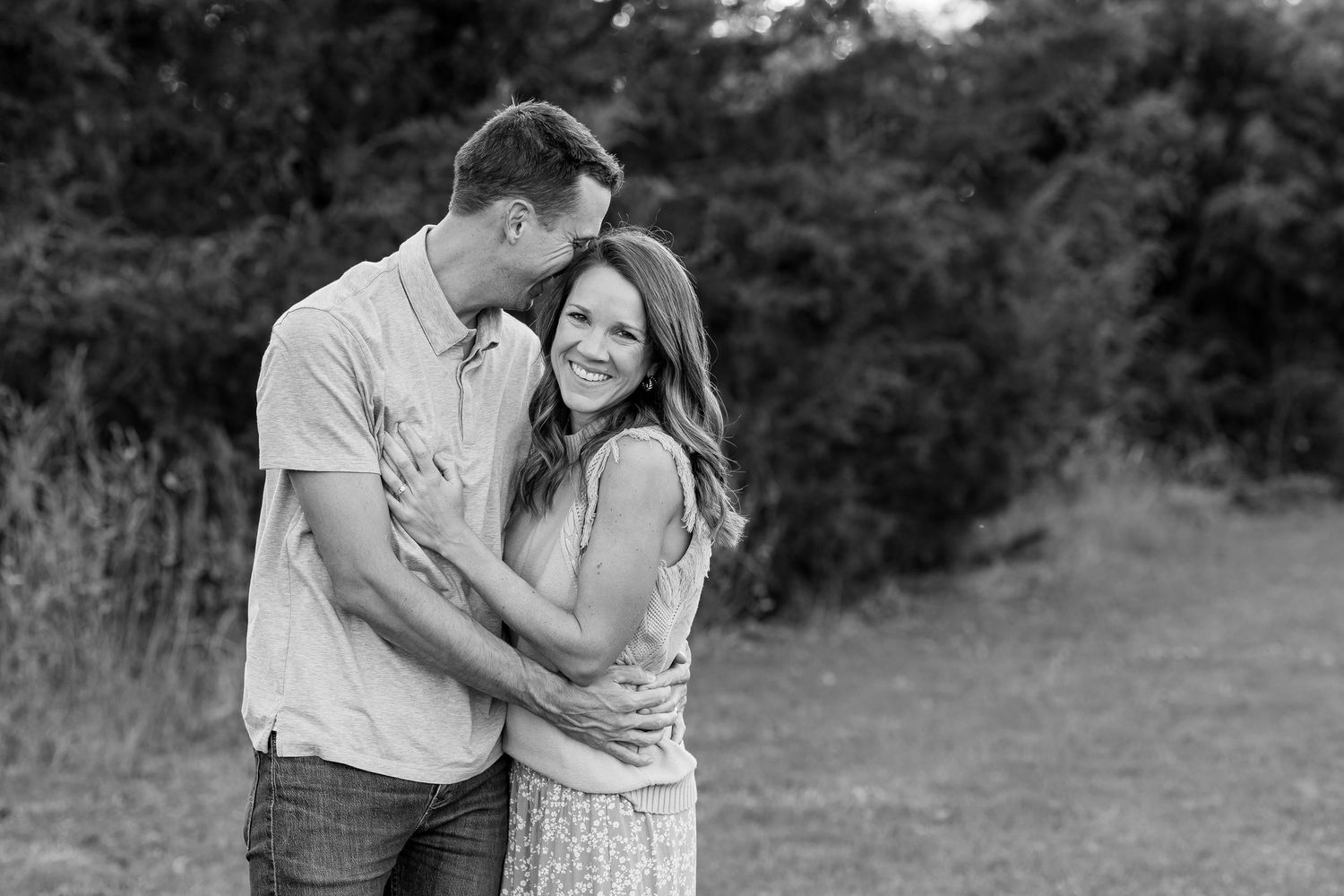 A couple embraces lovingly in a black and white outdoor portrait with a natural background.