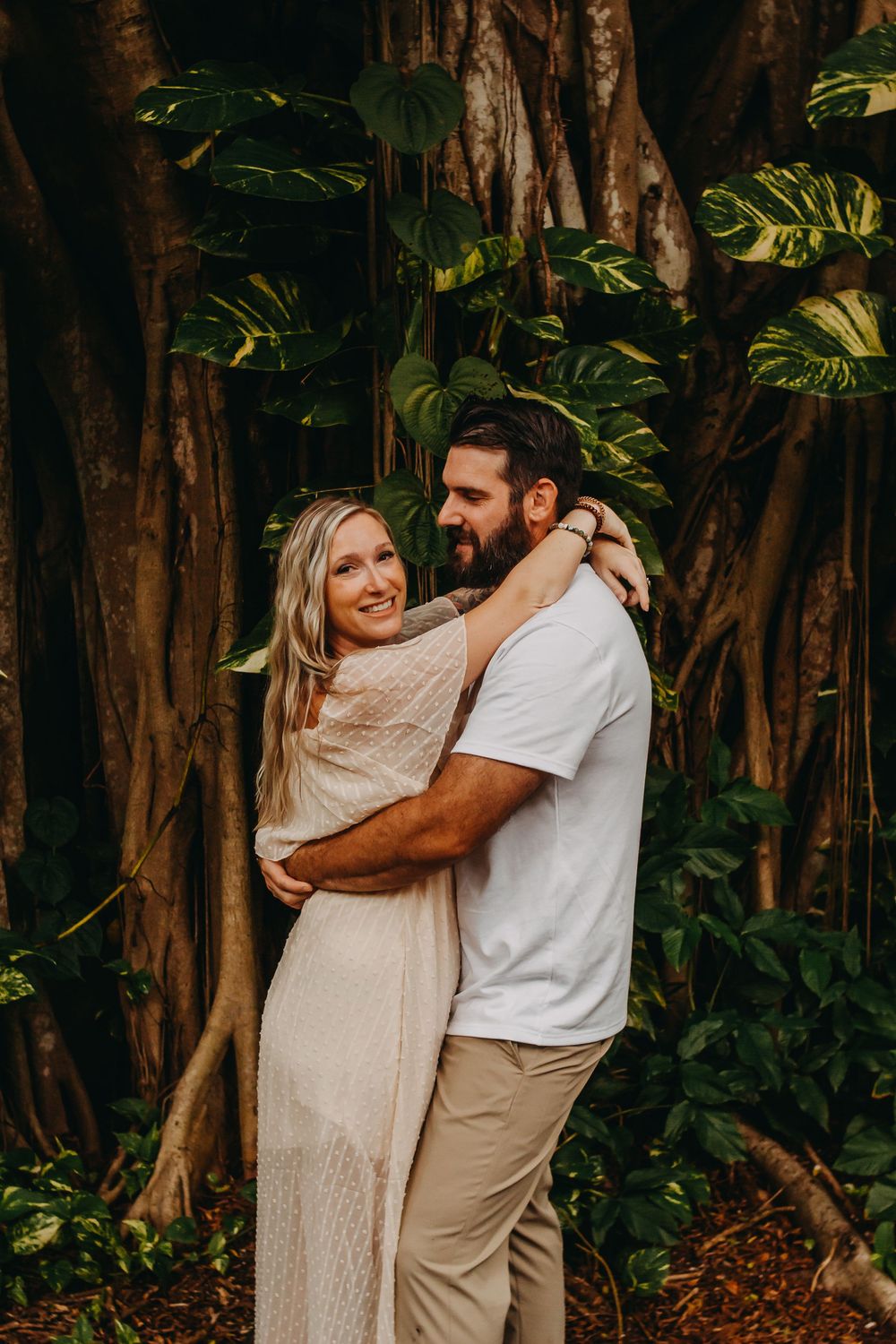 A couple embraces lovingly in a lush tropical garden setting with banyan trees and greenery in the background.