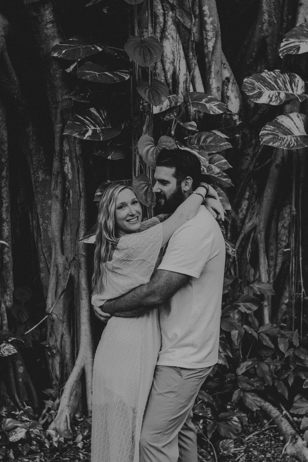 Black and white photo of a couple embracing in front of tropical foliage and tree trunks.