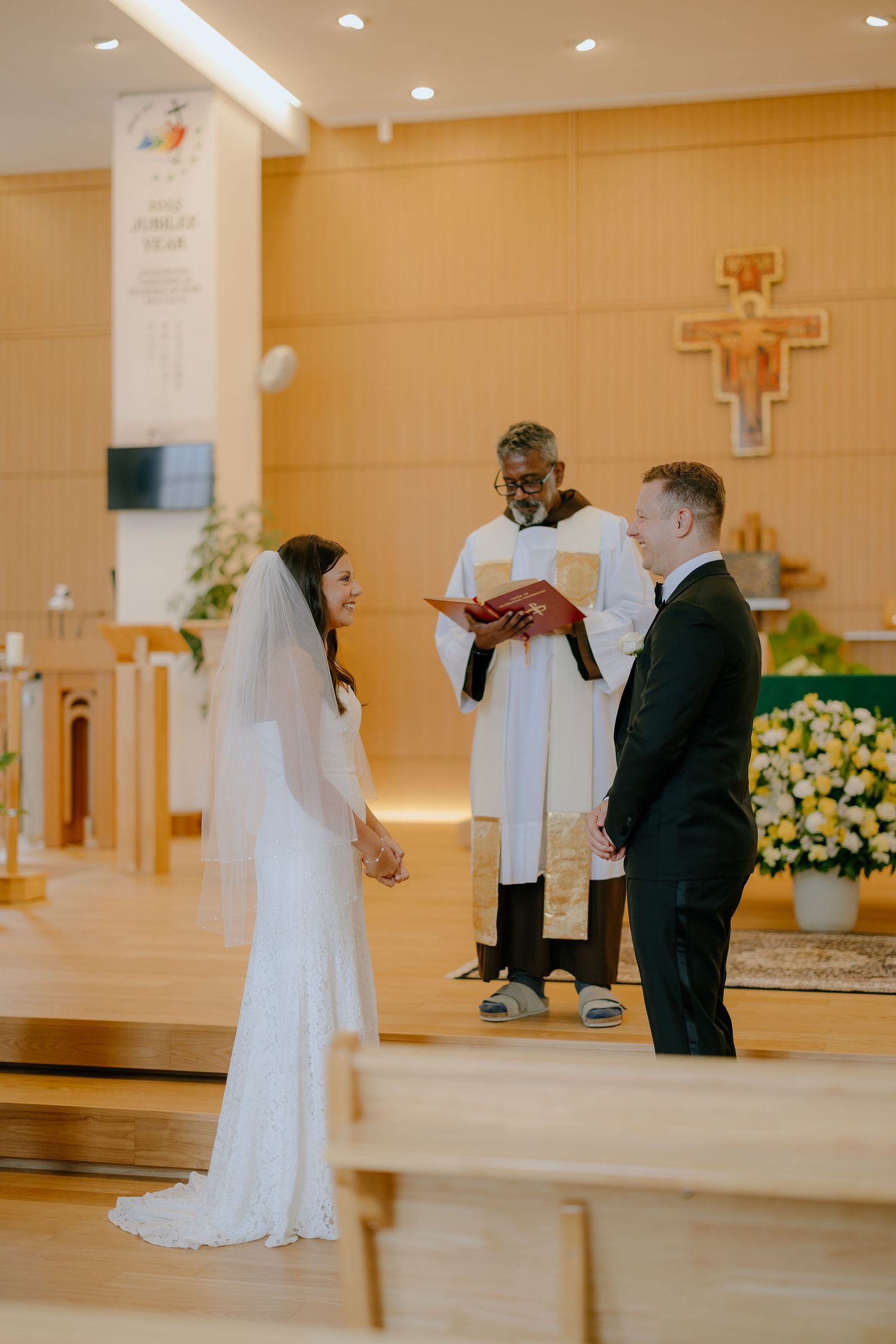 A wedding ceremony takes place in a modern church with warm lighting and a wooden cross on the wall.