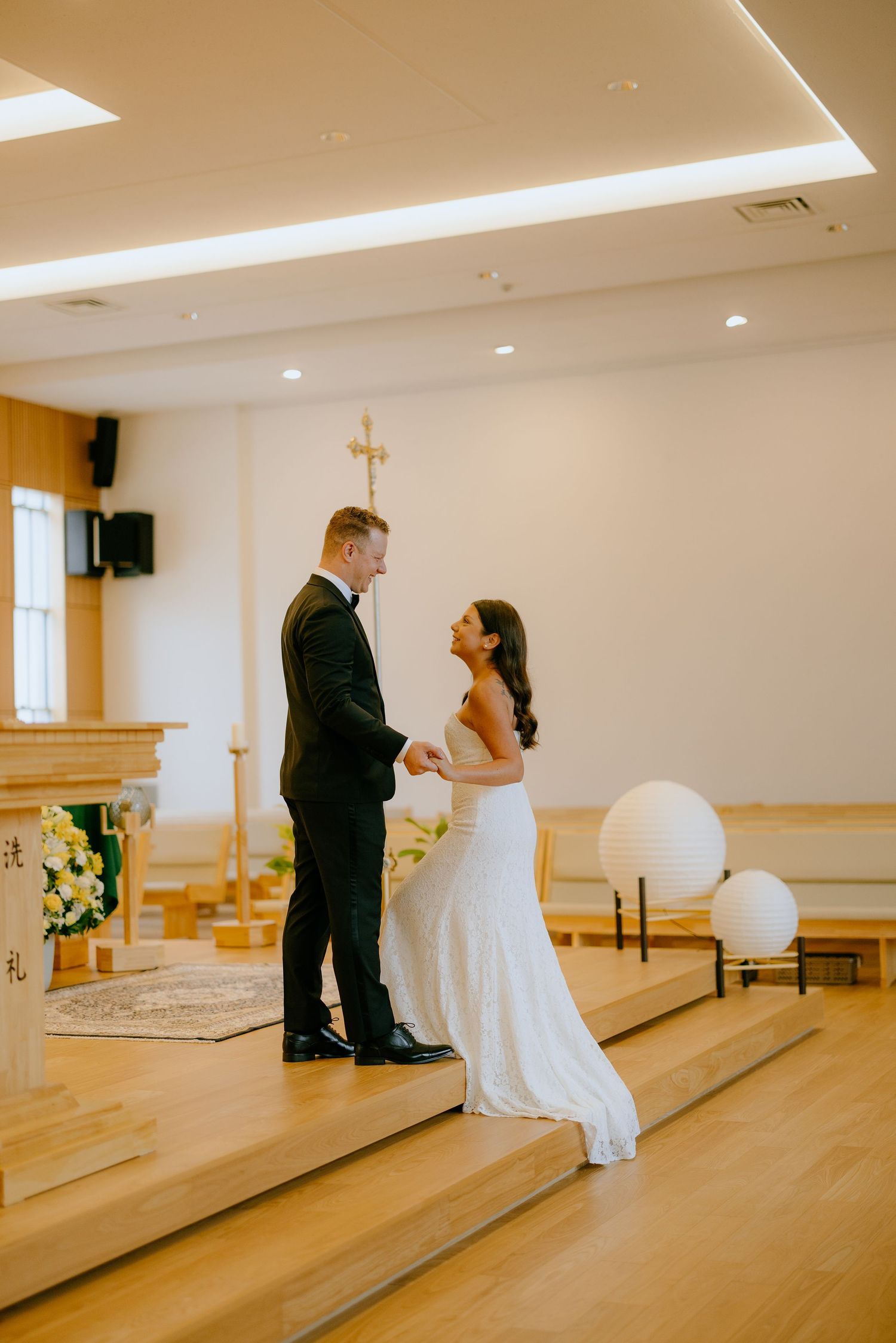 A couple in wedding attire share an intimate moment in a bright church sanctuary with wooden floors and white walls.