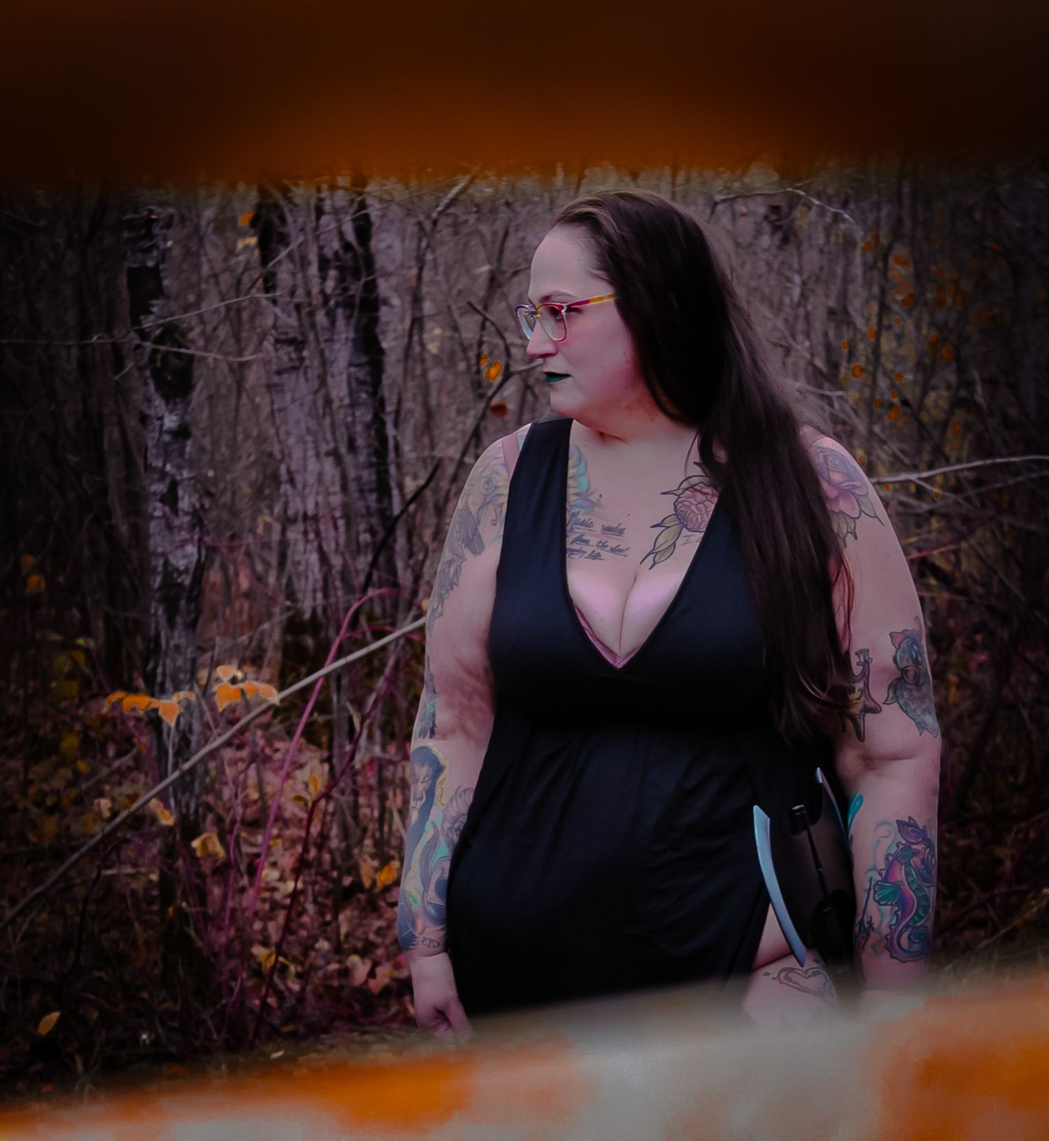A woman in a black sleeveless top poses outdoors with artistic tattoos visible against a dark wooded background.