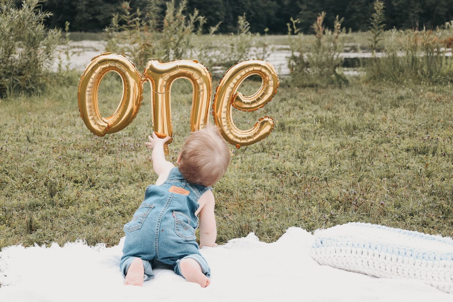 Baby sitting on blanket outdoors with gold letter balloons in background.