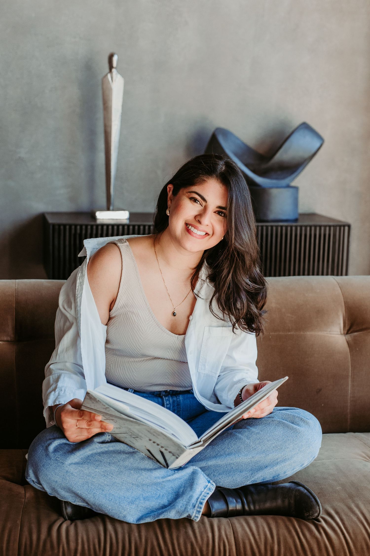 Casual lifestyle photo showing relaxed pose on brown leather couch with white cardigan and blue jeans in modern setting.