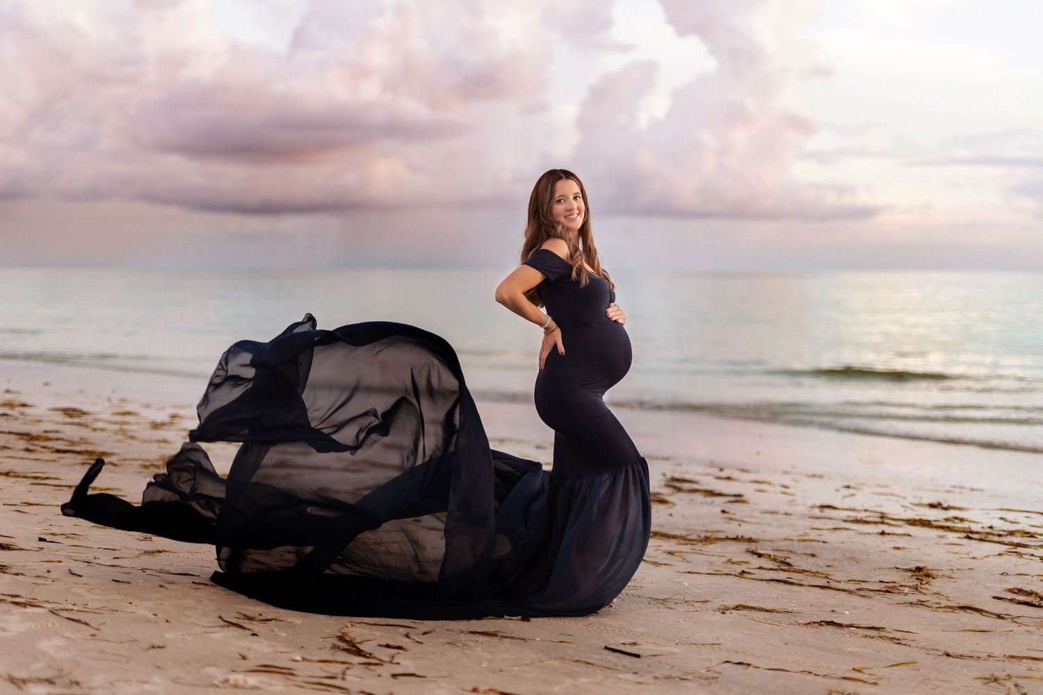 Dramatic maternity beach portraits with flowing navy dress at sunset.