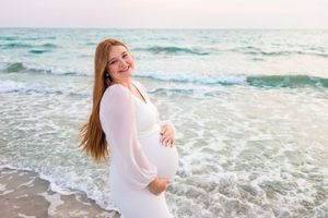 Ethereal beach maternity photoshoot with flowing white dress against turquoise waves and golden sunlight.