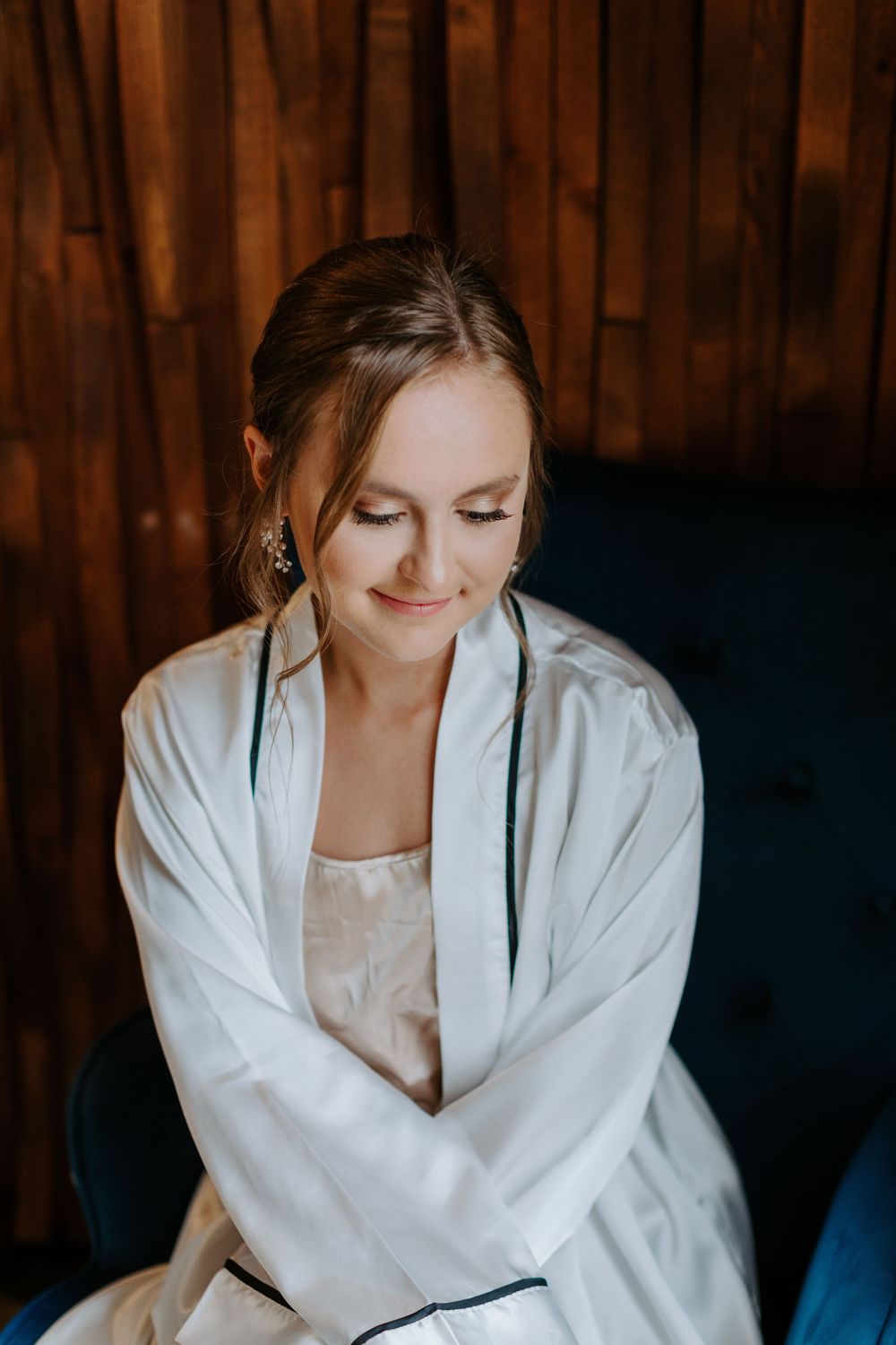 Person in white robe smiles gently while seated on blue chair with wood paneling background.