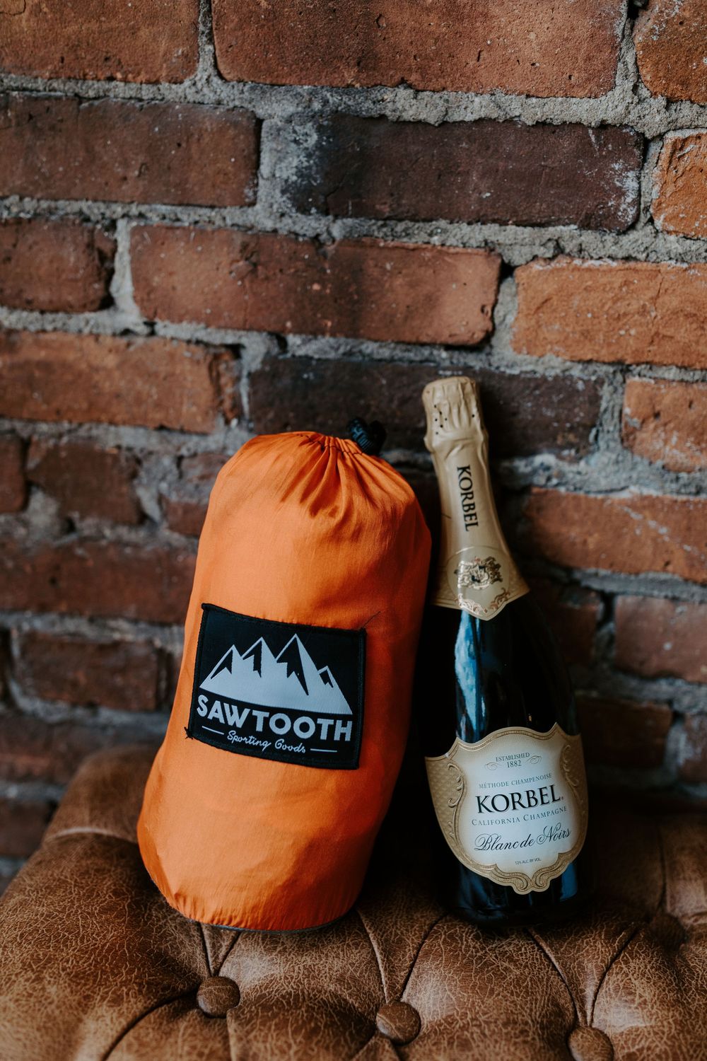 Orange camping gear and champagne bottle arranged against rustic brick wall backdrop.