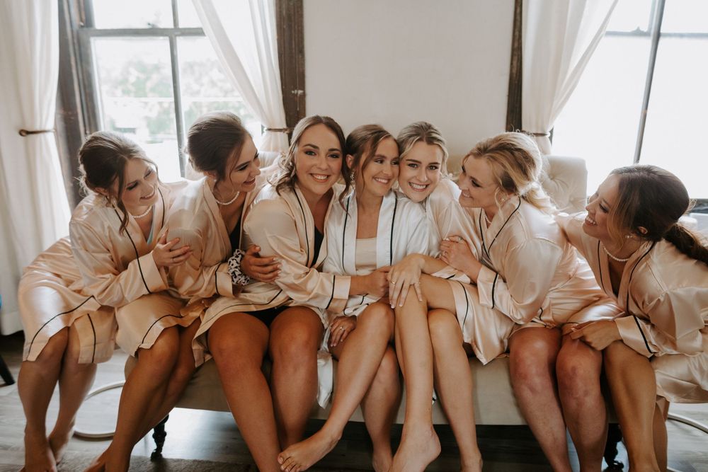Bridesmaids in matching robes sitting together on white couch during wedding preparations.