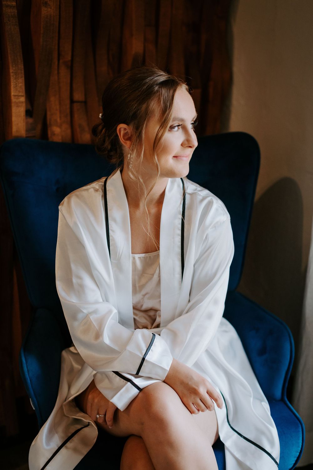 Person in white robe with black trim sits thoughtfully on blue velvet chair against wooden wall.