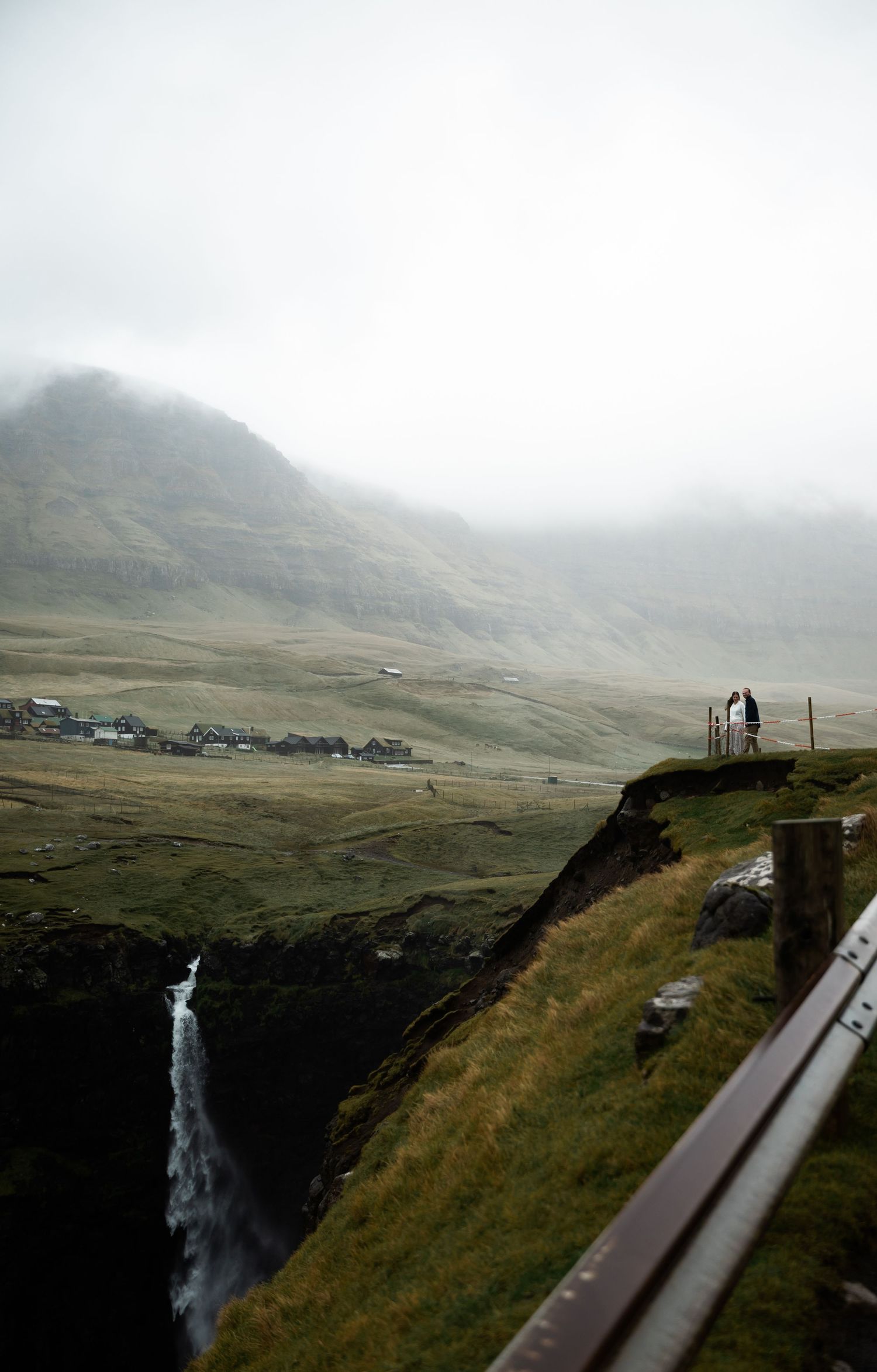 A scenic mountain road leads to a misty waterfall in the Faroe Islands on a moody, overcast day.