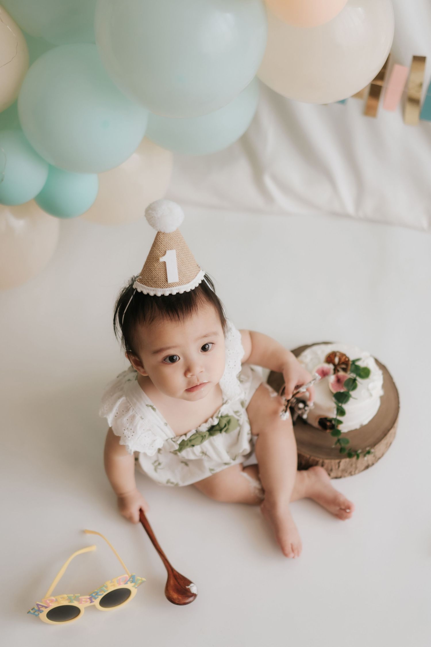 Baby in white dress lying on floor with mint green balloons and party decorations for first birthday celebration.