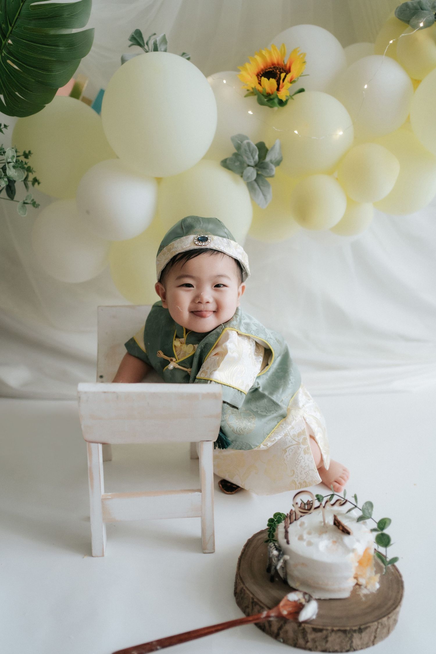 A festive birthday setup with white and yellow balloons, a sunflower accent, and a rustic cake display on a wooden slice.